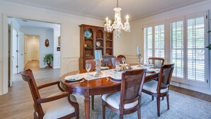 A dining room with a wooden table set for six, surrounded by six chairs, a chandelier, and a large window with blinds.