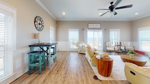 A spacious living room with a wooden floor, a ceiling fan, a dining area with a table and chairs, and a large window with blinds. - Gulf Front! Elevator! Just steps to the ocean! (Cape San Blas)
