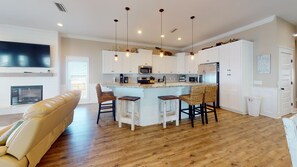 A modern kitchen with white cabinets, a central island with bar stools, a flat-screen TV above the island, and a fireplace in the living area. - Gulf Front! Elevator! Just steps to the ocean! (Cape San Blas)