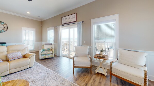 A spacious living room with a beige sofa, two armchairs, a coffee table, and a rug. There are large windows with blinds, a sliding glass door leading to a balcony, and a clock on the wall.
