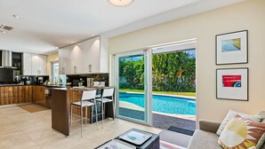Una cucina moderna con mobili bianchi, un'area pranzo con vista piscina e un soggiorno con divano e cuscini.