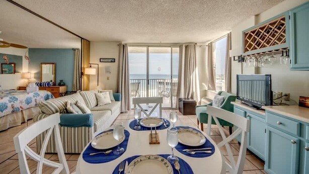 A room with a dining table set for four, a sofa, a TV, and a view of the sea.