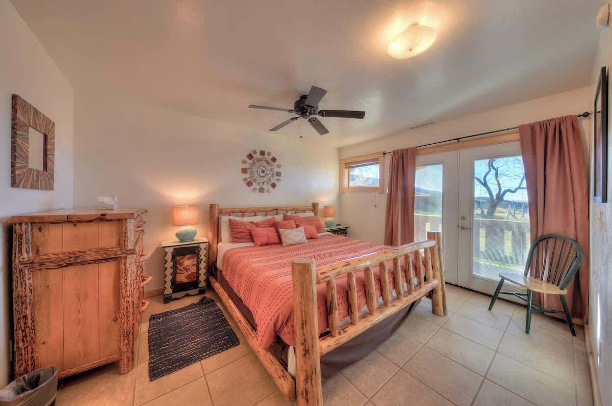A bedroom with a wooden bed, a ceiling fan, a window with curtains, and a chair by the window.