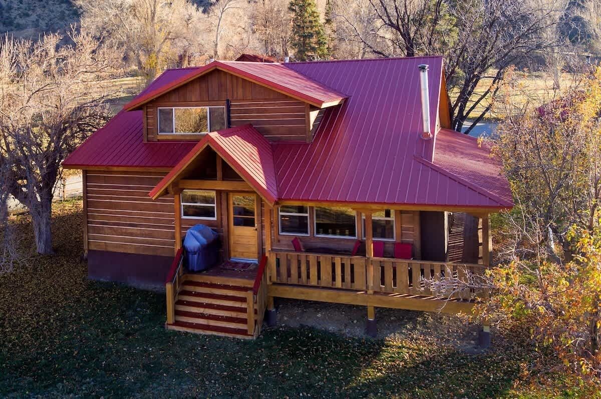 A two-story wooden cabin with a red roof, a porch with wooden railings, and a small staircase leading to the entrance.