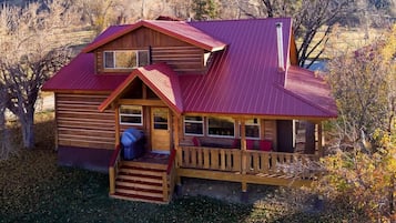 A two-story wooden cabin with a red roof, a porch with wooden railings, and a small staircase leading to the entrance.