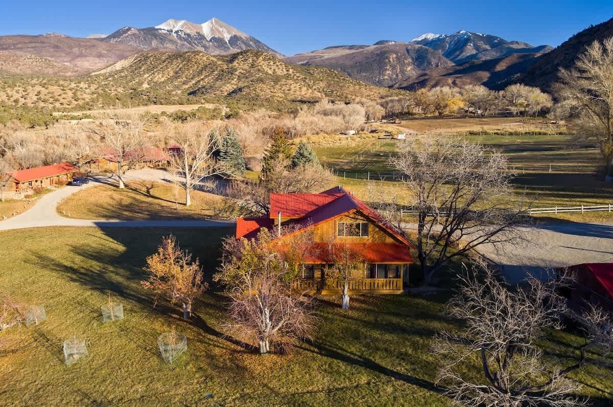 A rural landscape with a house featuring a red roof, surrounded by trees and a mountain range in the background.