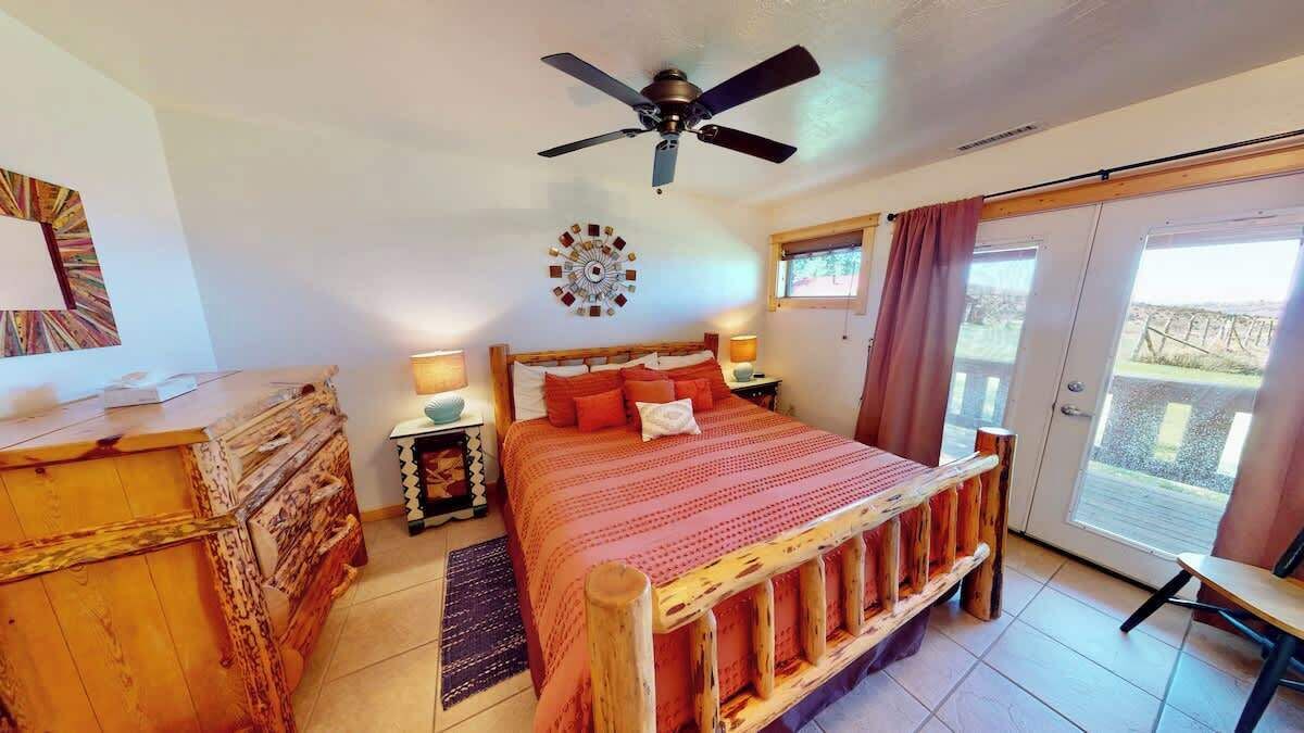 Quarto com cama, mesas de cabeceira, ventilador de teto, janela com cortinas e vista para o exterior.
