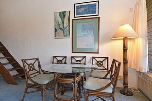 A dining area with a round glass table surrounded by wicker chairs, a framed painting above the table, and a floor lamp to the right.