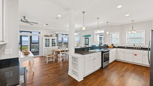 A modern kitchen with white cabinetry, a black countertop, and a large island. There is a dining area with a table and chairs, and a view of the ocean through large windows. - 5 Bedroom Beachfront (Cape San Blas)