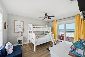 A room with a bunk bed, a desk, a chair, a small table, a lamp, a wall decoration, and a view of the beach.