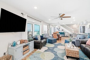 A spacious living room with a large flat-screen TV, comfortable seating, a staircase leading to an upper level, and a view of the outdoors.
