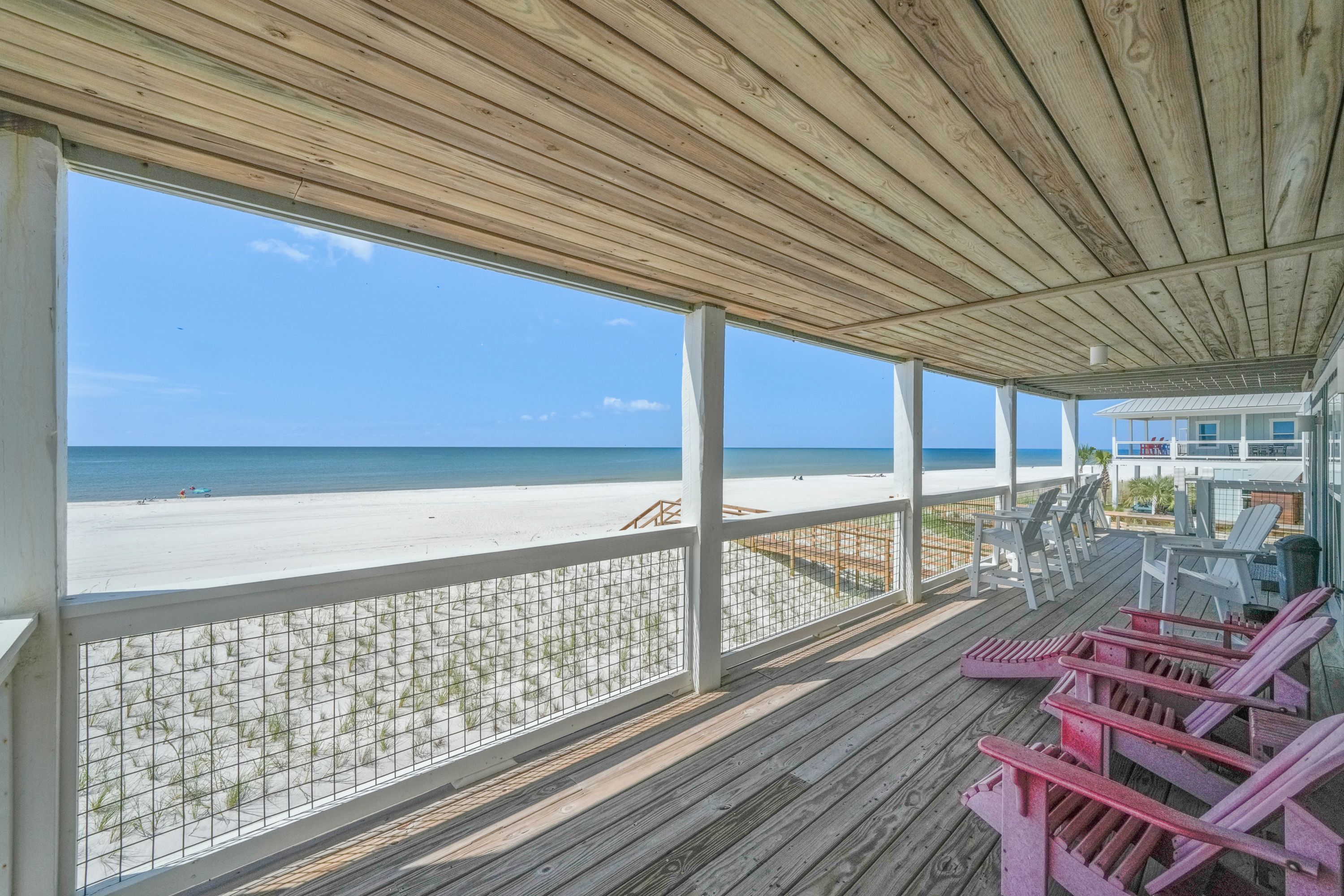 A beachside wooden deck with a view of the ocean, featuring a railing and several Adirondack chairs.