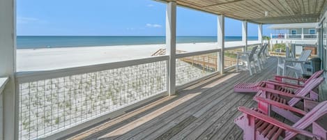 A beachside wooden deck with a view of the ocean, featuring a railing and several Adirondack chairs.