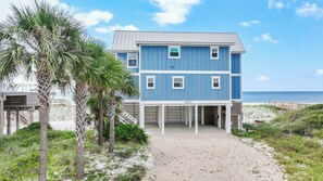 A two-story beachfront property with a covered porch and a gravel driveway leading to the entrance.