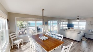 A spacious living room with a wooden dining table, white chairs, a grey sofa, and a television. Large windows offer a view of the ocean. - Pet Friendly Beach Front Home (Cape San Blas)