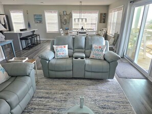 Una sala de estar moderna con un sofá de cuero gris, dos sillones a juego y una mesa de comedor con sillas. Hay almohadones decorativos con motivos playeros en el sofá.