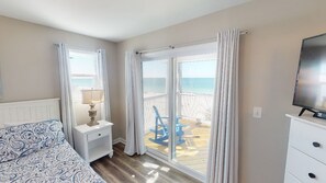 A bedroom with a bed, nightstand, lamp, and a TV. There is a balcony with a chair and a view of the beach.
