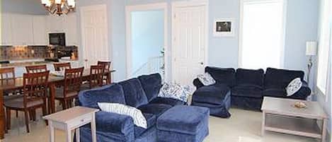 A living room with a blue sectional sofa, a dining area with wooden chairs, and a kitchen in the background.