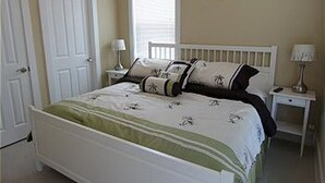 Et soveværelse med en hvid seng, et natbord med lampe, en dør og et vindue med persienner.