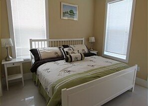 A bedroom with a white bed, two bedside tables with lamps, a window with blinds, and a framed picture on the wall. - 1st Tier Home with Private Pool! (Cape San Blas)