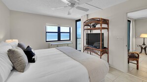 A bedroom with a large bed, a ceiling fan, a TV on a wooden stand, and a view of the sea through a window.