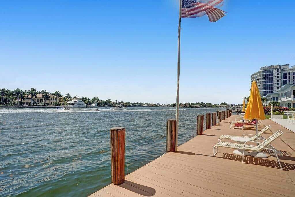 A wooden pier with lounge chairs and a yellow umbrella, overlooking a body of water with buildings and palm trees in the background.