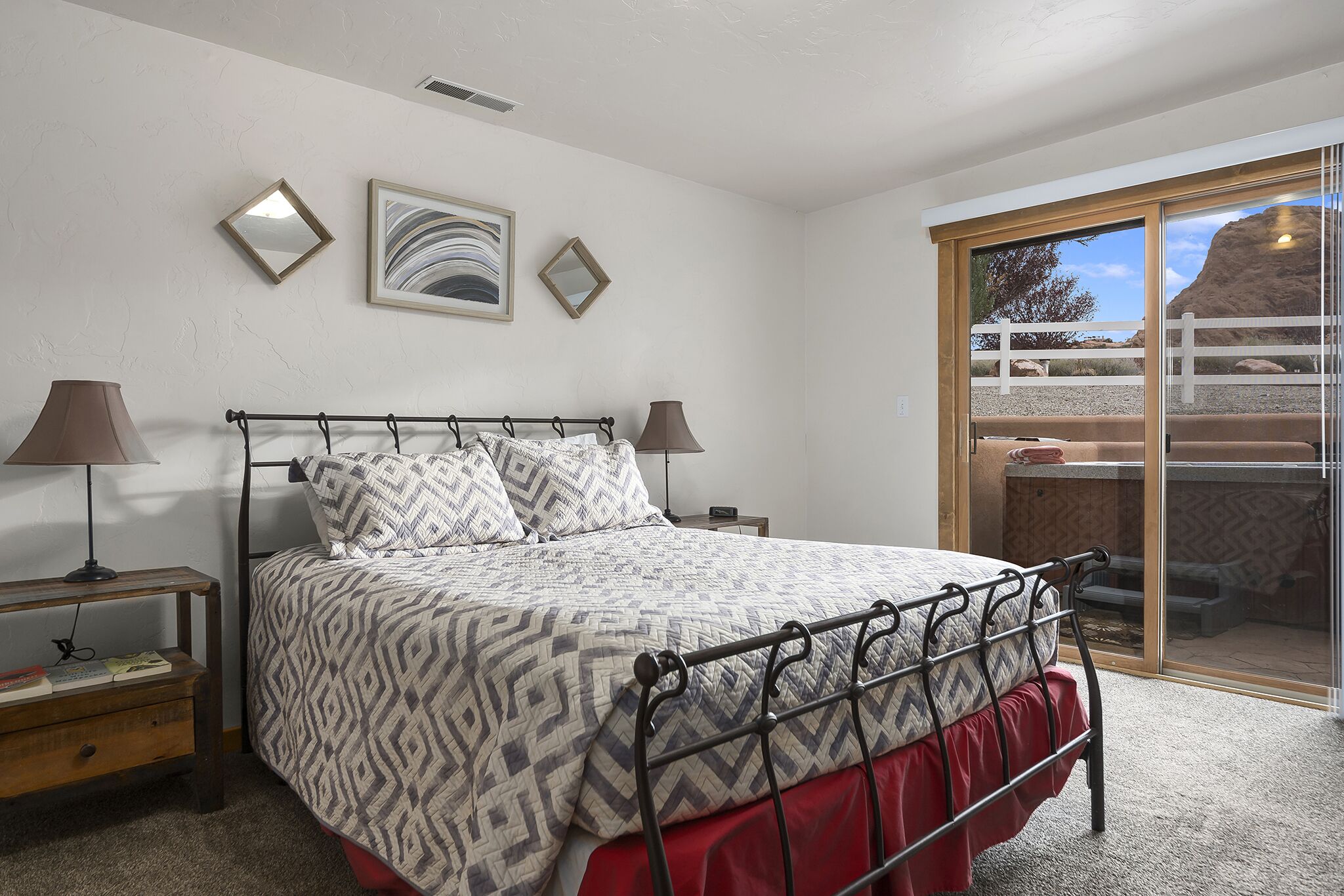 A bedroom with a bed, two bedside tables, a mirror, and a view of a desert landscape through a sliding glass door.