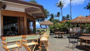 An outdoor seating area with wooden tables and chairs, thatched umbrellas, and a view of palm trees and the ocean.