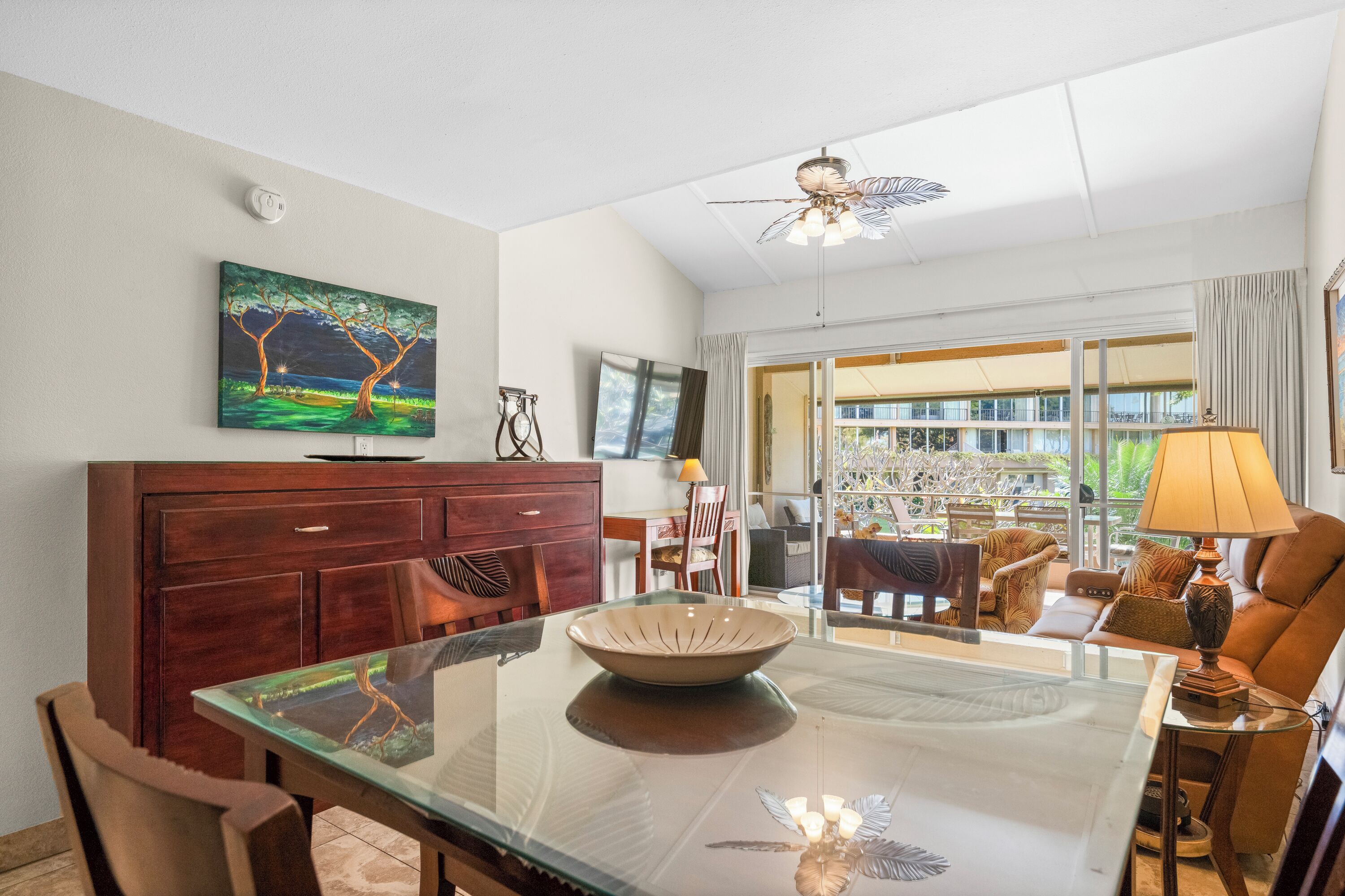 A modern dining area with a glass table, wooden chairs, a wooden cabinet, a painting, a ceiling fan, and a view of a balcony.