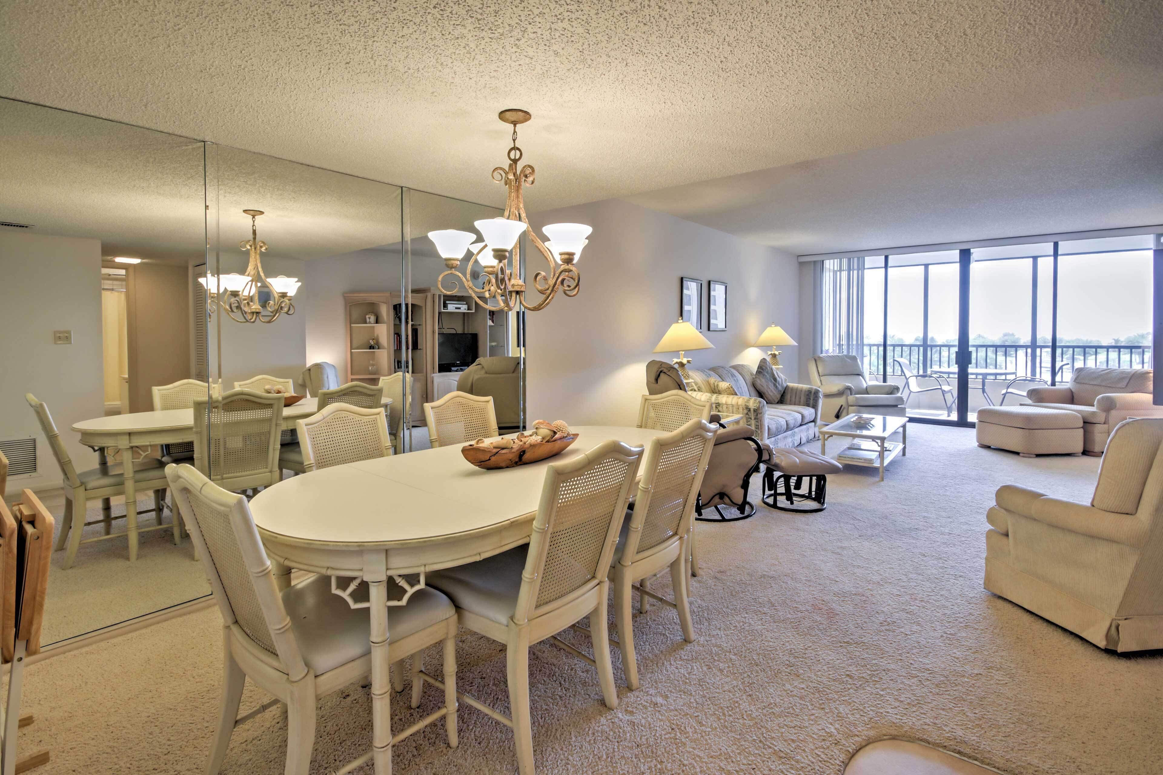 A spacious living room with a large round table, several chairs, a sofa set, and a balcony with a view.