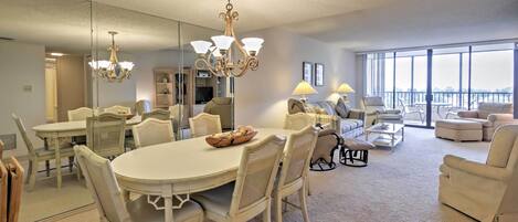 A spacious living room with a large round table, several chairs, a sofa set, and a balcony with a view.