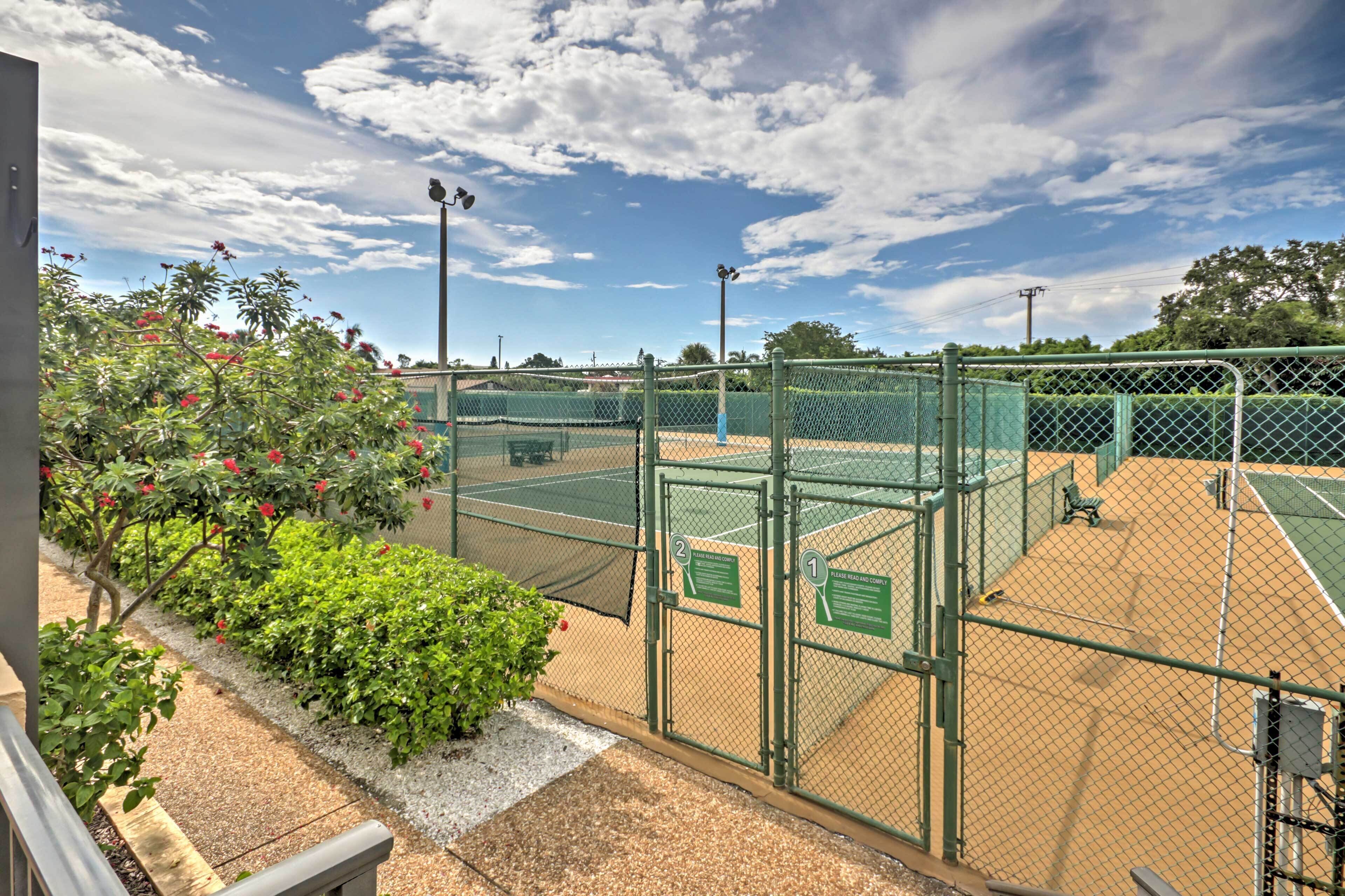A tennis court with a chain-link fence, a bench, and a small garden with red flowers.