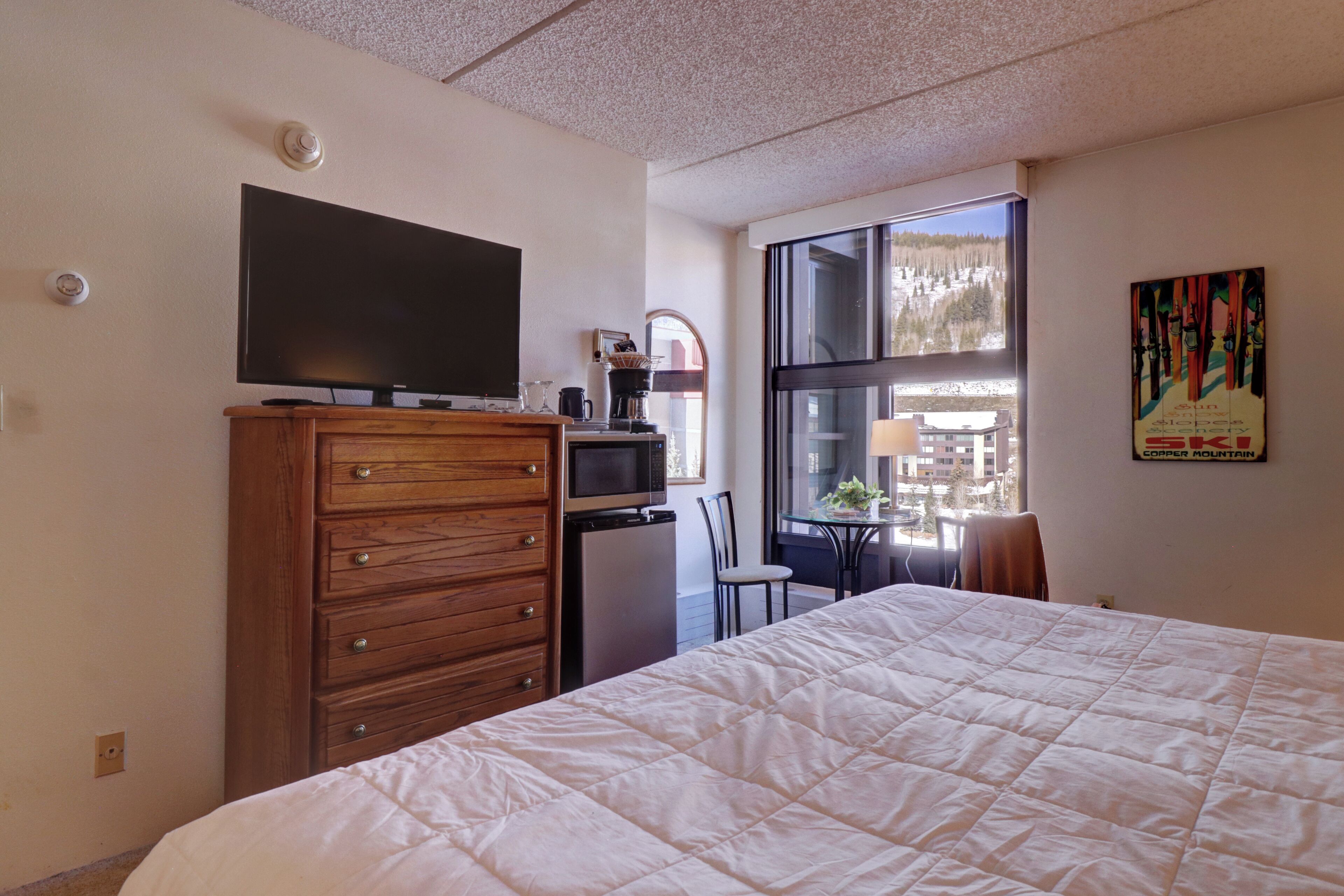 A hotel room with a bed, a dresser, a television, a small refrigerator, a window with a view of trees, and a table with chairs.