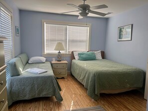 Une chambre avec deux lits simples, un ventilateur de plafond, une table de chevet avec une lampe et une fenêtre avec des stores.