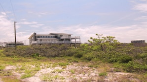 A two-story beachfront house with a balcony, surrounded by dense vegetation and sandy terrain.