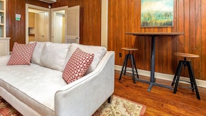 A room with a white sofa, two red patterned pillows, a wooden wall, a painting, and two stools. - Frogtown Cottage | Historic Downtown Savannah, Pool, Pet Friendly (Savannah)