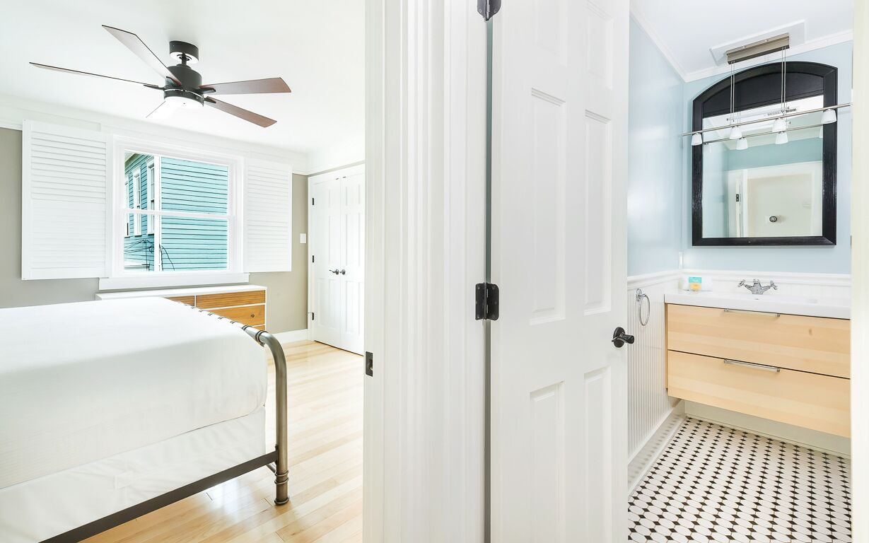 A bedroom with a bed, a ceiling fan, a window with blinds, a door, and a bathroom with a sink and mirror.