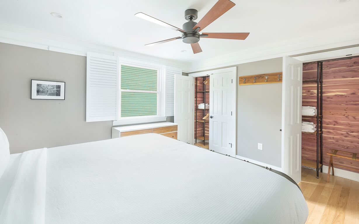A bedroom with a large bed, a ceiling fan, a window with blinds, and a wooden closet.