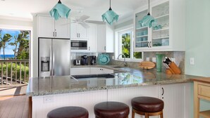A modern kitchen with white cabinets, a stainless steel refrigerator, a microwave, and a coffee maker. There are three hanging pendant lights, a large island with stools, and a view of the ocean through the window. - Poipu Palms #201 By Parrish Kauai- oceanfront views & see turtles! (Koloa)