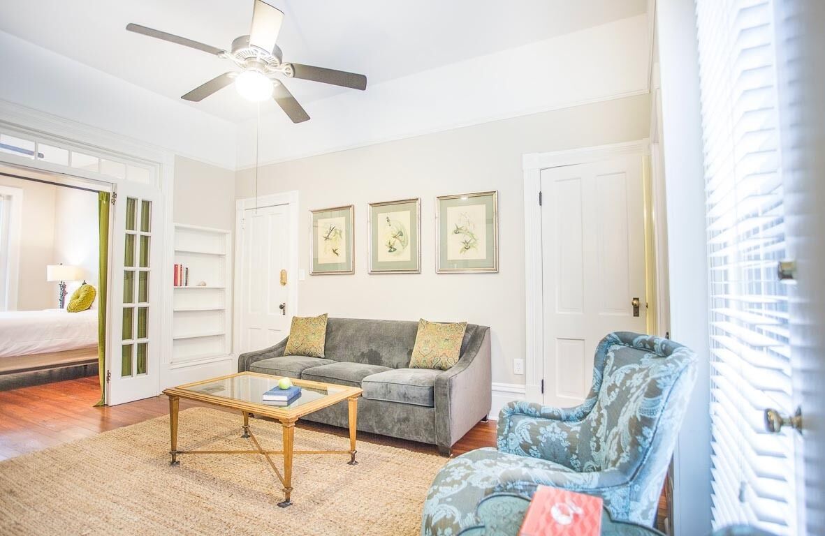 A living room with a grey sofa, a blue armchair, a coffee table, and a ceiling fan. There are framed pictures on the wall and a door with a green curtain.