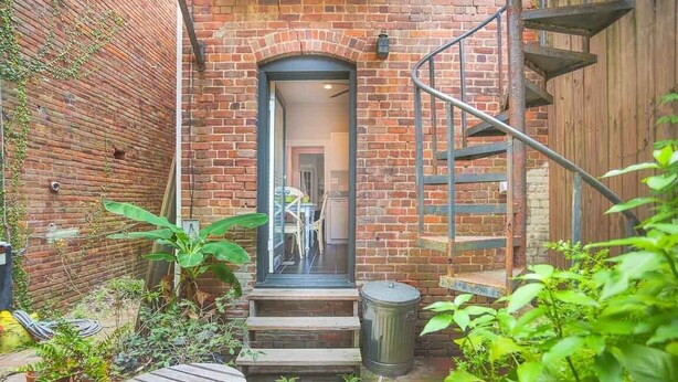 A brick building with a staircase leading to a door, surrounded by greenery and a small patio area. - Well Appointed Row Home w/ King, Across from Forsyth Park by Lucky Savannah (Savannah)