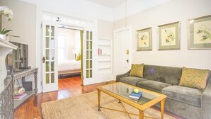 A living room with a sofa, a coffee table, a television, and a door leading to another room. - Well Appointed Row Home w/ King, Across from Forsyth Park by Lucky Savannah (Savannah)