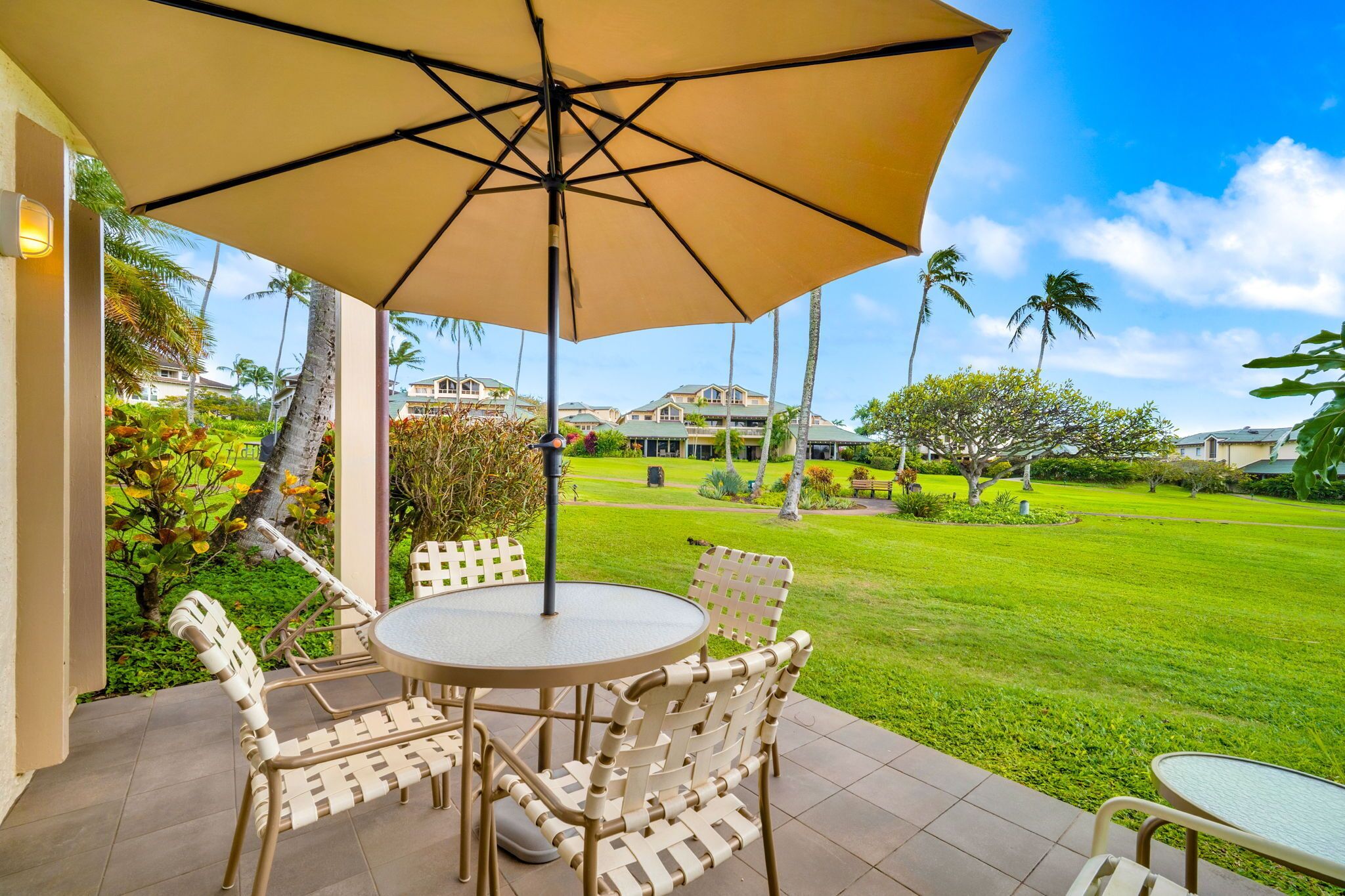 Une terrasse avec une table ronde et des chaises, un grand parasol, et une vue sur une pelouse bien entretenue et des bâtiments en arrière-plan.