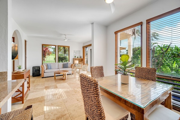Amplia sala de estar con mesa de vidrio, sillas de mimbre, un sofá y una mesa de centro. Ventanas grandes con persianas ofrecen vistas a la vegetación exterior.