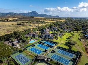 Vista aérea de un resort con múltiples canchas de tenis, una piscina y montañas al fondo.