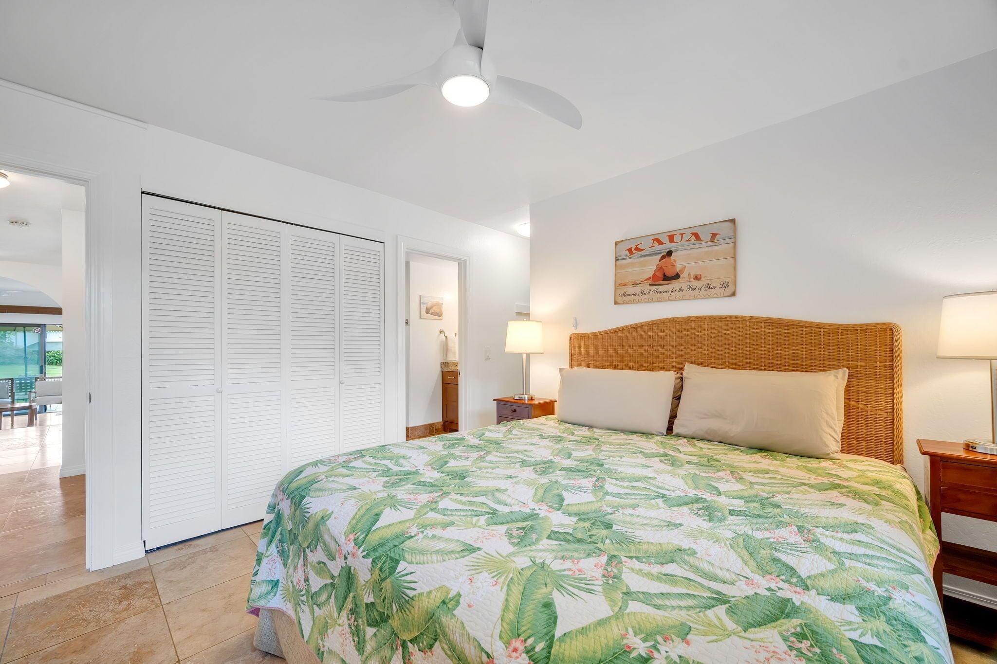 Un dormitorio con cama, ventilador de techo, mesita de noche, lámpara, cabecero de mimbre y una puerta que da a un balcón.