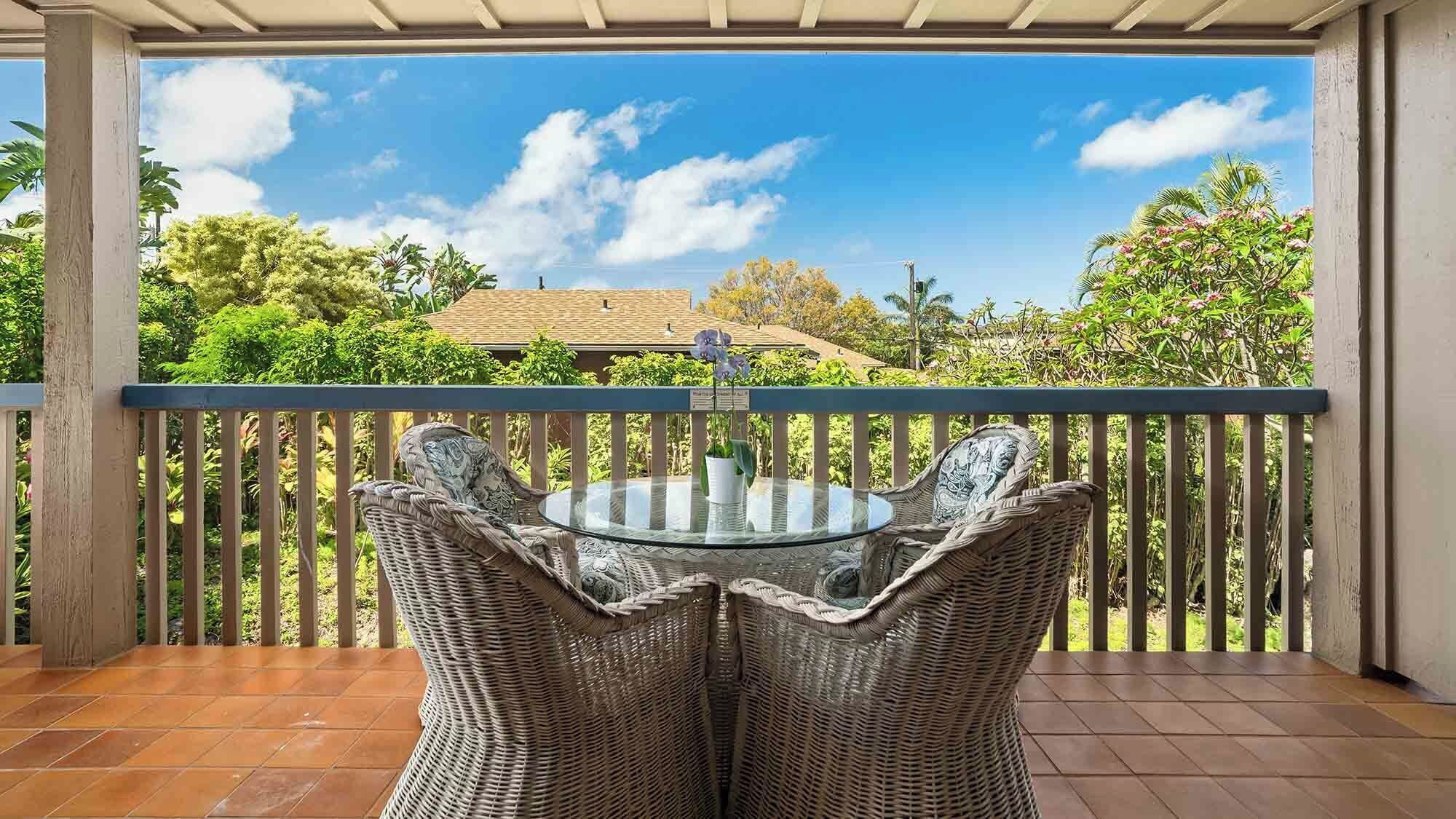 A covered balcony with wicker chairs and a glass table, overlooking a lush garden and a house.