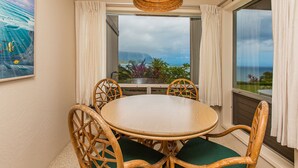 A round table with four chairs, a woven pendant light, and a view of the ocean through a large window with curtains.