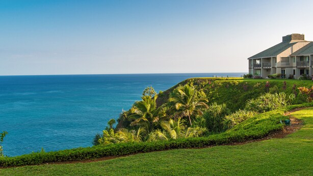 A coastal hotel with a view of the ocean, lush greenery, and a well-maintained lawn.