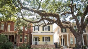 Una casa a due piani con un portico, un albero grande di fronte e un edificio in mattoni alla sinistra.